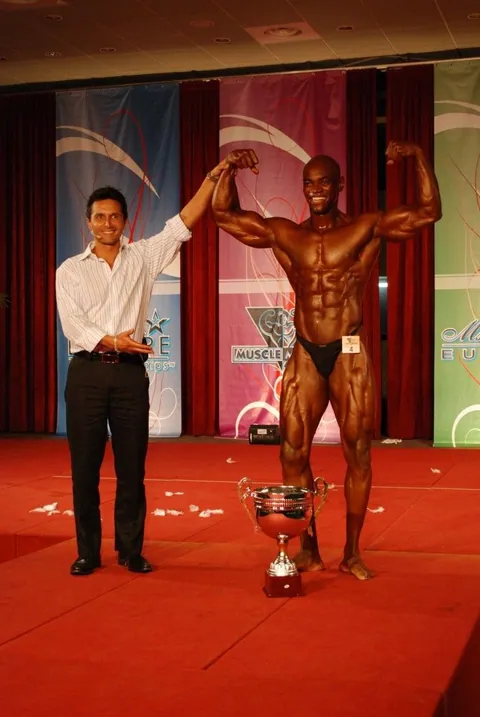 Antony Bessala bras leve sur scene a cote d'un grand trophee sous une banniere Musclemania Europe