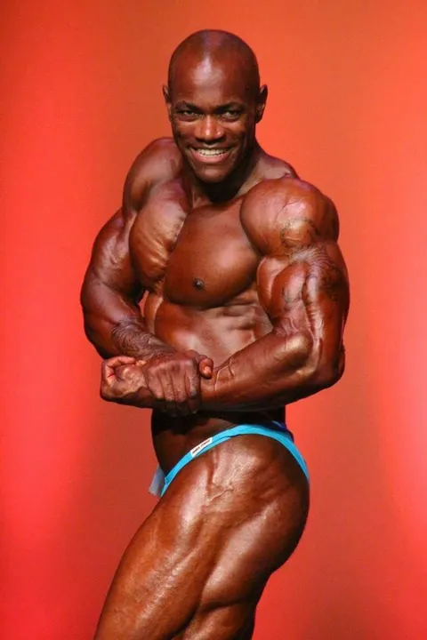 Antony Bessala sur la scene Musclemania Pro : pose de force en slip bleu sur fond orange